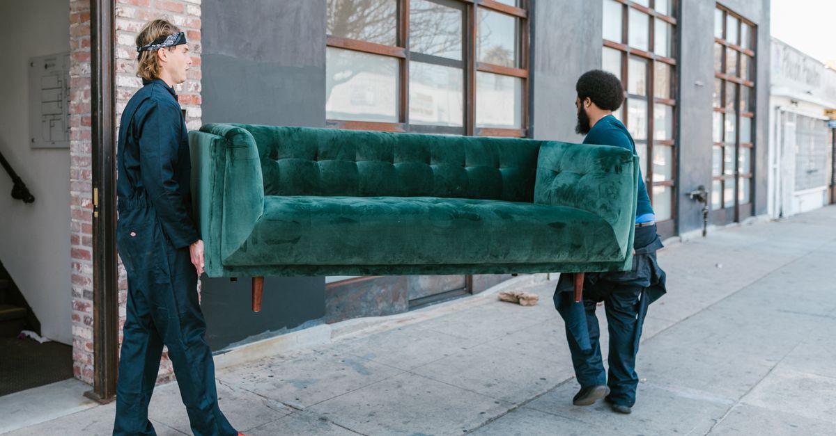 Zwei Männer tragen gemeinsam ein grünes Sofa über eine Straße in der Stadt.