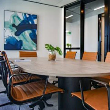 Interior view of an office room of a cleaning company. A large bright table with orange tilt chairs. The wall to the hallway is made of glass and on the wall hangs an abstract picture in different shades of blue