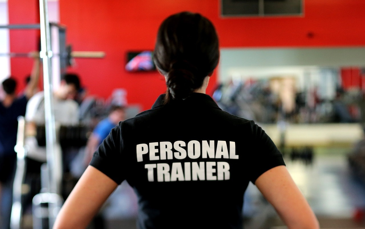 Ein Frau von hinten mit einem schwarzen T-shirt auf dem Personal Trainer steht in einem Fintess studio.
